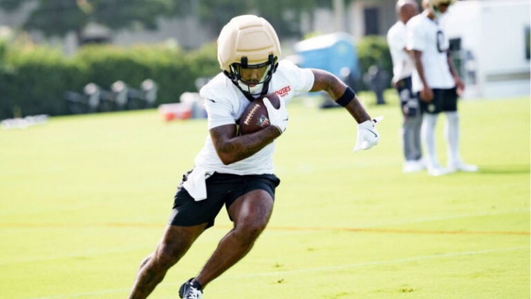A football player, possibly a Saints wide receiver, runs with the ball on a grassy field as teammates watch in the background.