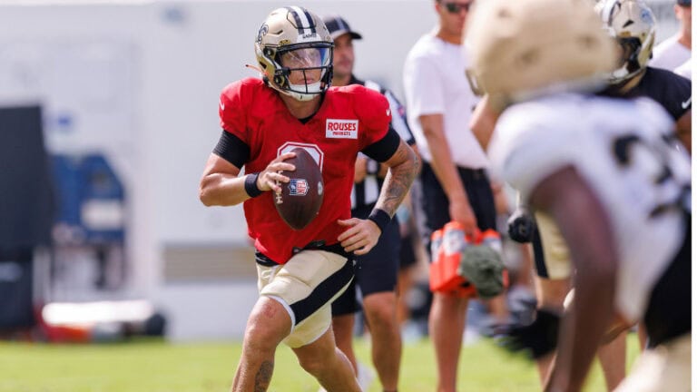 A football player in a red Jaguars jersey runs with the ball at practice as teammates watch position battles in the background.