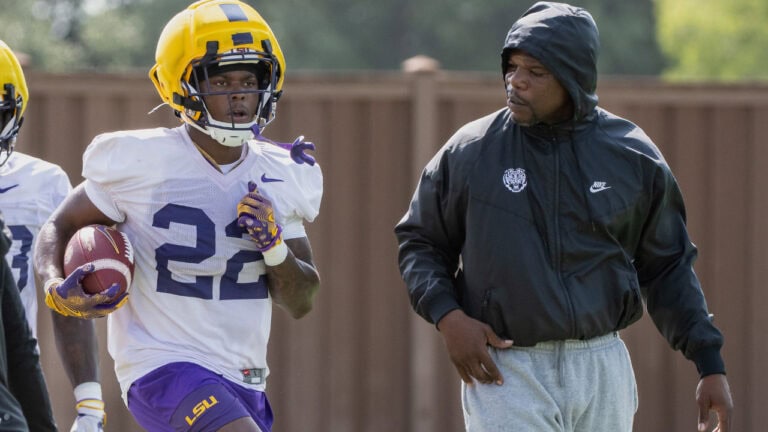 An LSU football player runs with the ball at practice while a coach in a black Nike hoodie watches closely.