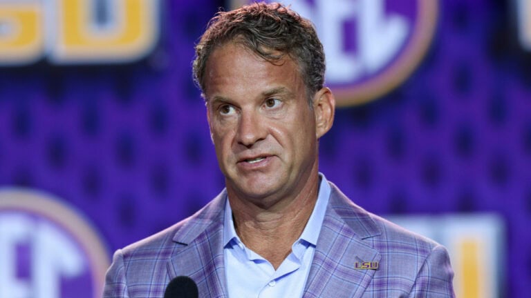 A man in a plaid suit jacket speaks at a microphone in front of a purple LSU backdrop with SEC logos.