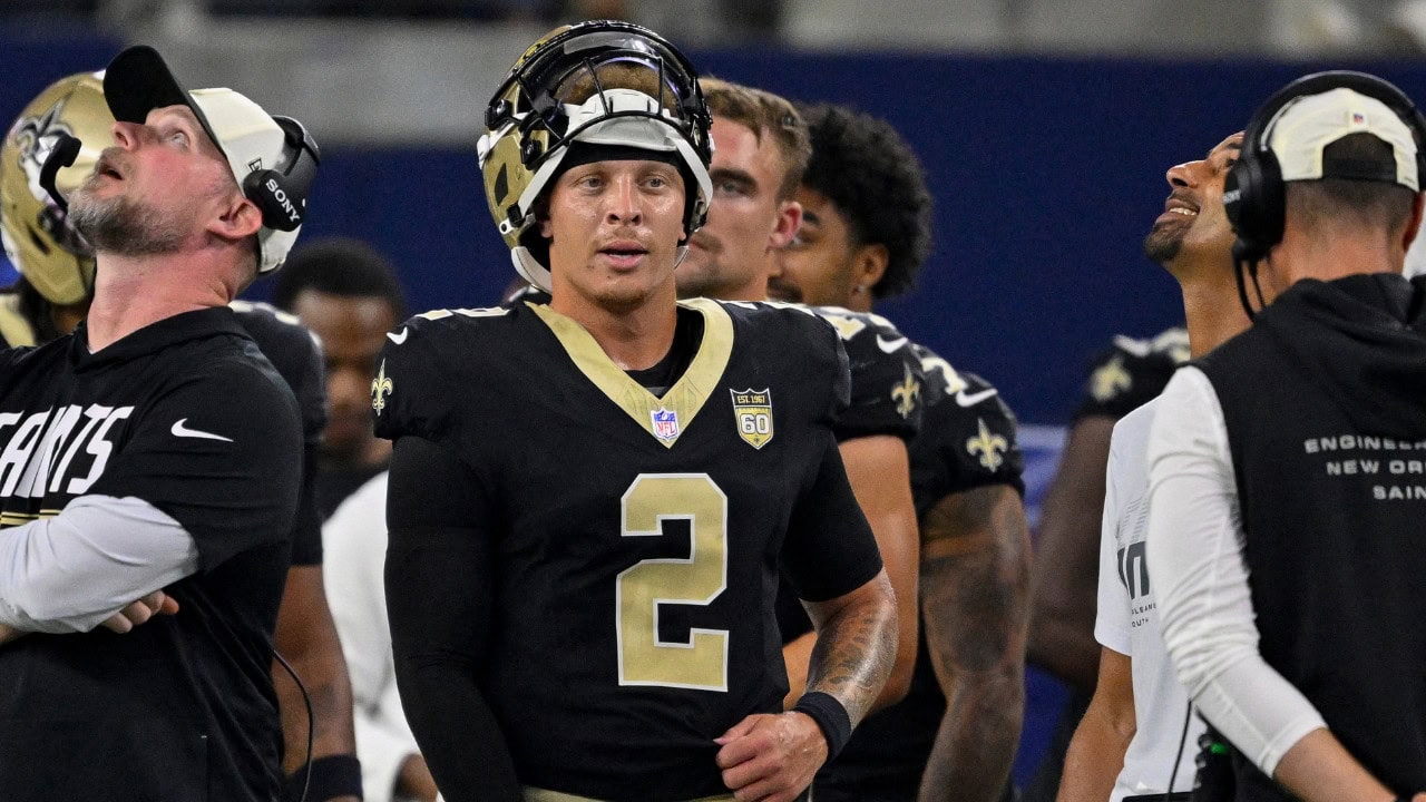 Spencer Rattler, wearing a black No. 2 Saints jersey, stands on the sideline with coaches and teammates.