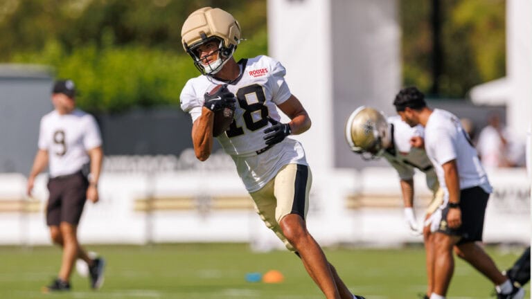 A football player in a white jersey runs with the ball at practice, while teammates and a coach watch from the green field in the background.