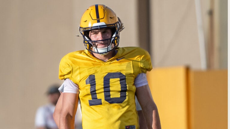 Smiling football player in a yellow number 10 jersey stands outdoors during an LSU scrimmage, glancing slightly to the side.