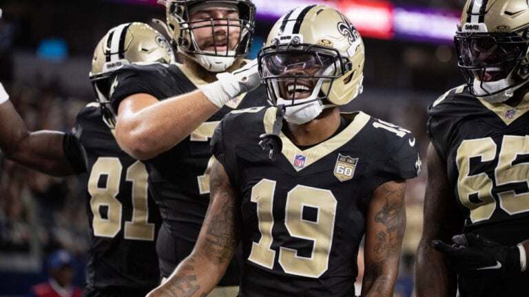 Four New Orleans Saints players stand on the field, with the center player smiling.