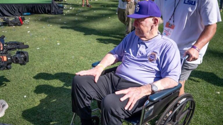 An older man in a wheelchair wearing an LSU hat and polo sits on grass while Skip Bertman stands behind him.