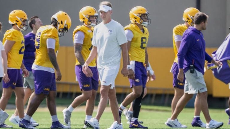 LSU football players in yellow helmets and purple shorts walk with coaches in white and purple gear during practice.