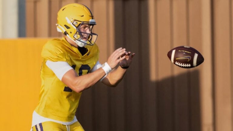 An LSU player in a yellow uniform gets ready to catch a football during fall camp, with a brown wall behind him.