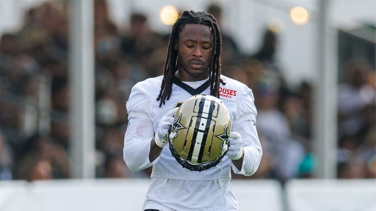 Saints player in a white jersey holds a gold helmet at practice, fans blurred in the background.