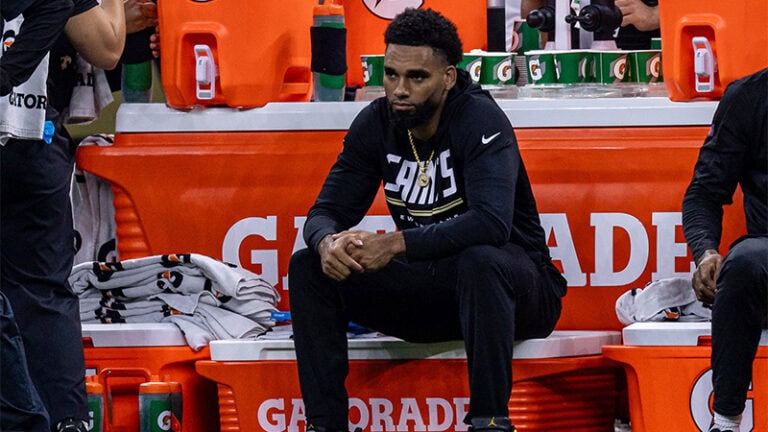 Saints player Olave sits on the bench in black, gazing ahead seriously as the team’s injury report casts a shadow over him.