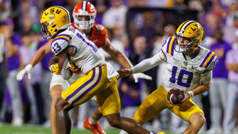 An LSU football player hands off the ball during a game against a team in orange uniforms.