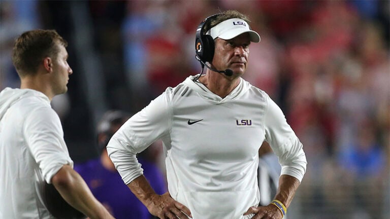 An LSU coach stands on the sideline with a headset and hands on hips, appearing dejected, next to another coach in a white shirt.