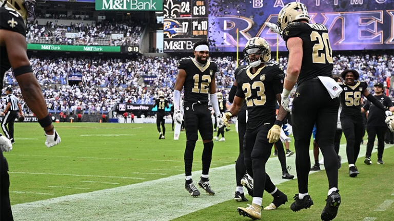 Football players in black uniforms huddle as a “Baltimore Ravens” sign glows in the background; can the Saints claim victory?.
