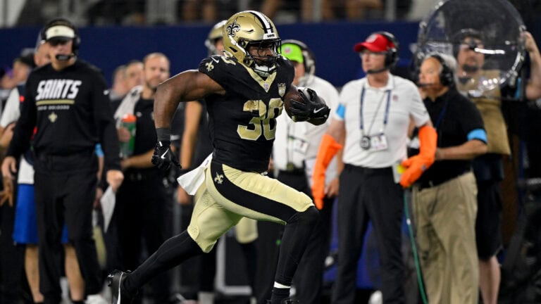 A Saints player in black and gold runs with the football as coaches and staff watch from the Week 3 sidelines.