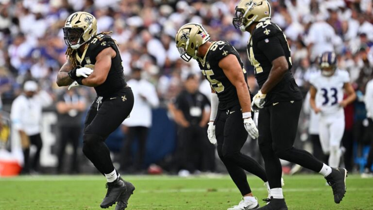Three New Orleans Saints players in uniform play on the field as a cheering crowd watches from the stands.