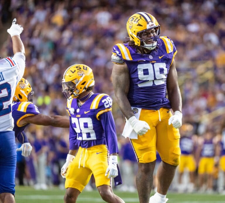 Three LSU football players in purple and gold uniforms stand on the field during a stadium game.