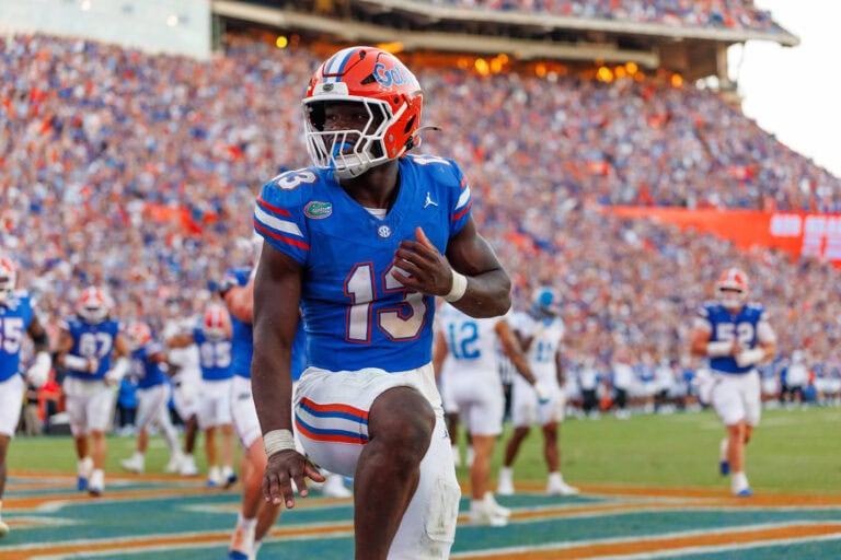 An LSU football player in a blue jersey runs across the field as the crowd and fellow players cheer in the background.