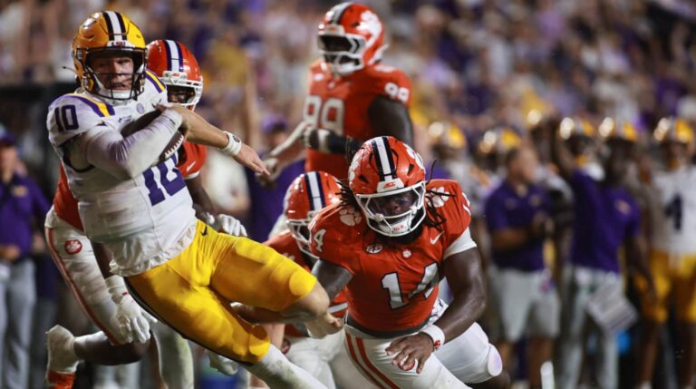 During a game, a player in yellow and purple is tackled mid-air by an opponent wearing orange.