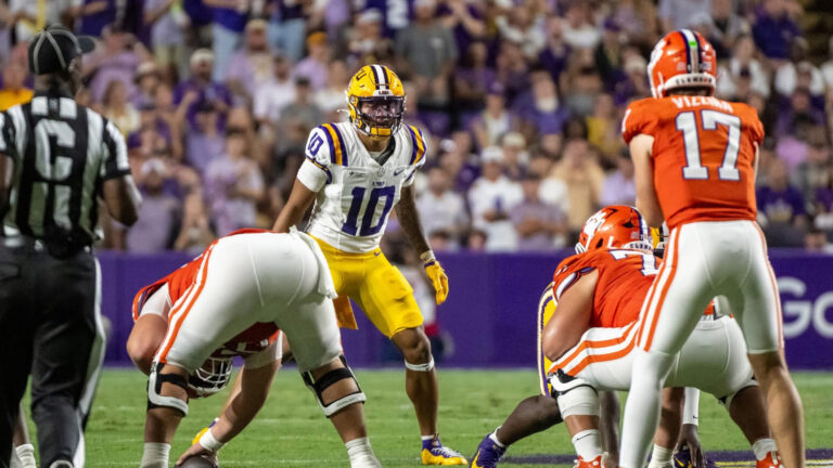 LSU and Clemson players line up for a play as a referee watches nearby, with fans filling the stands in the background.