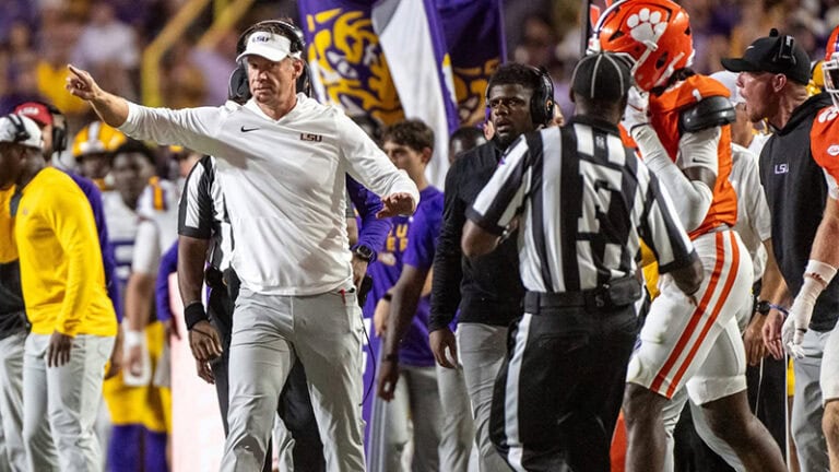 An LSU coach gestures on the football sideline as players, coaches, and referees stand nearby during the game.