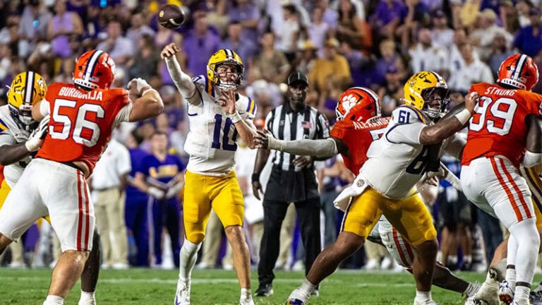A quarterback in white and yellow throws a football as orange-clad defenders close in during a game.