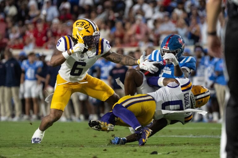 LSU players tackle an Ole Miss opponent in a college game as the crowd and Lane Kiffin’s team watch the action unfold.
