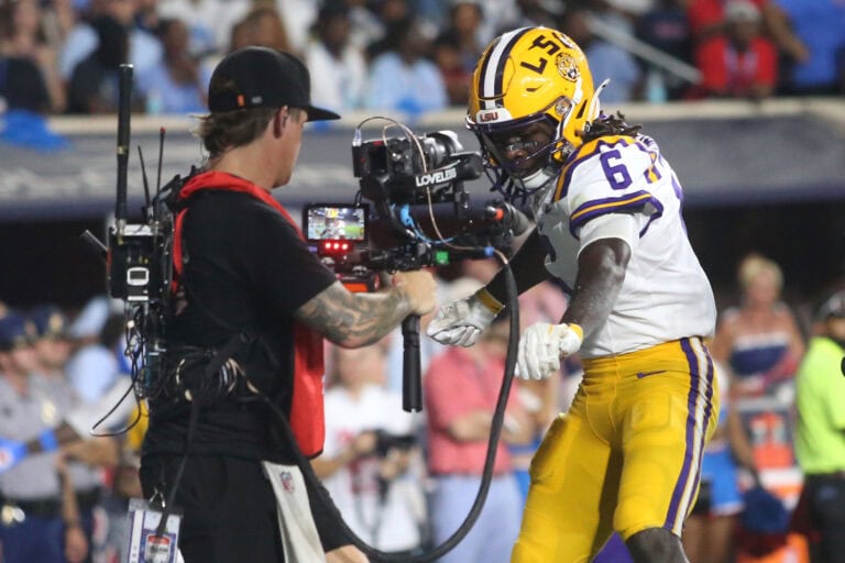 An LSU football player in a yellow and white uniform poses for a cameraman on the field as fans watch in the background.