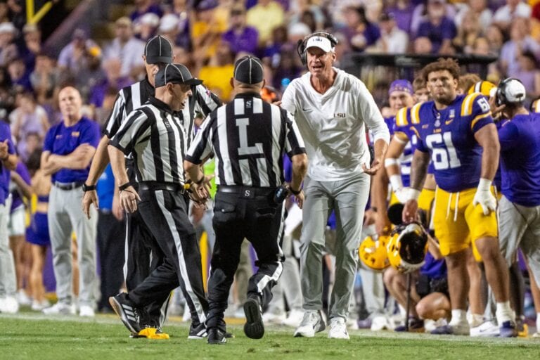 A football coach in white yells at referees as players and fans look on during a tense Southeastern Conference game.