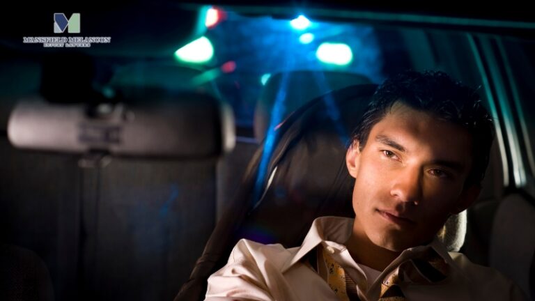 A man sits in a car at night as flashing police lights behind him light up his face and the car’s interior.