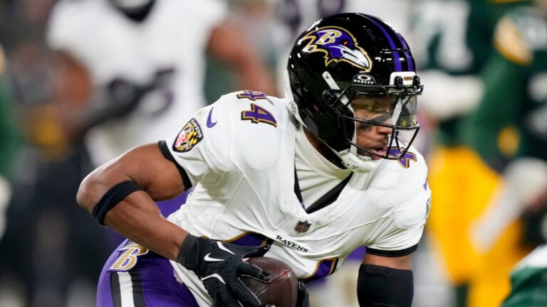 A Baltimore Ravens player in a white uniform runs with the ball as opponents pursue him on the field.