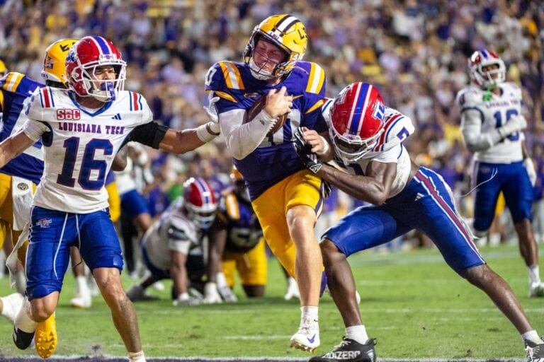An LSU player in purple and yellow runs as two opponents in white, blue, and red move in to tackle him on the field.