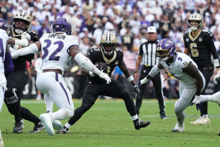 Travis Etienne Jr., in black, carries the ball as two Saints defenders in white try to tackle him on the grass field.