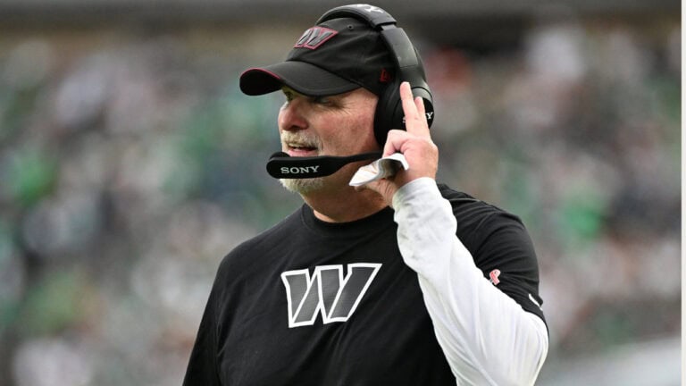 Dan Quinn, NFL coach in a black “W” shirt and headset, stands on the field during a game.