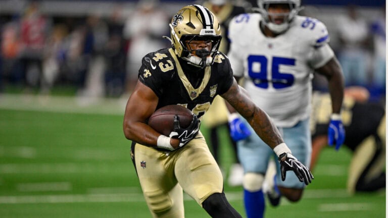 A Saints player in black and gold runs the football as opponents in white chase him down the field.