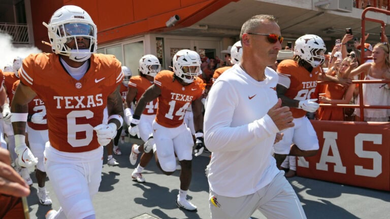 Texas Longhorns football players and their coach jog onto the field as cheering fans take photos, capturing the excitement of game day.