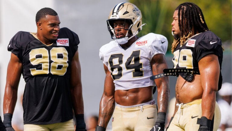 Three Saints edge rushers in black and gold uniforms gather outdoors at a 2026 season practice.