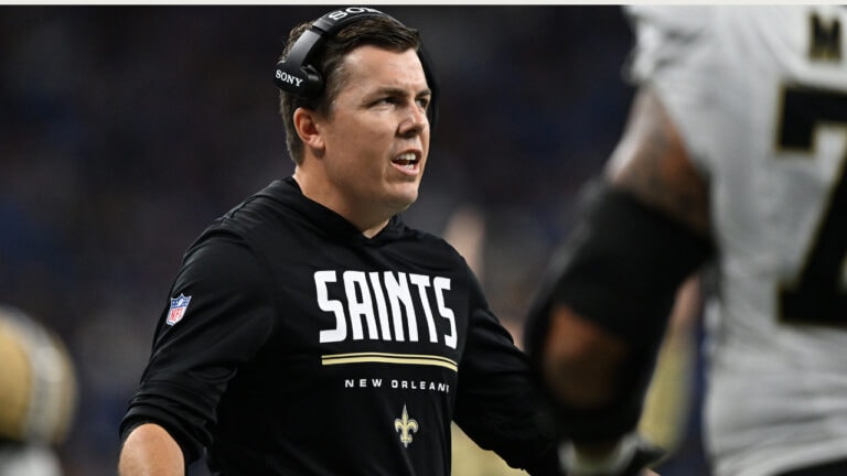 A football coach in a black “Saints New Orleans” sweatshirt watches the sideline, focused on a potential comeback during the game.