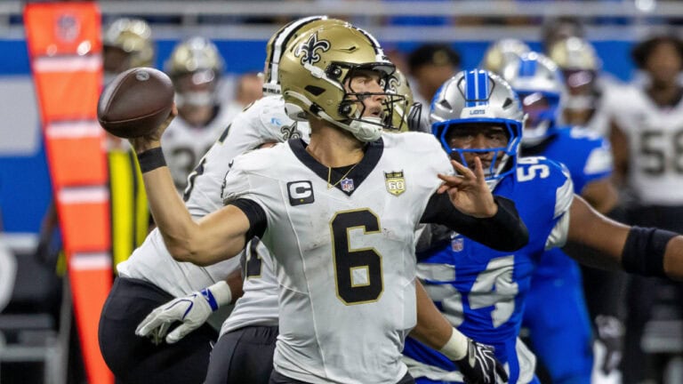 Saints QB Tyler Shough, in a white uniform, prepares to throw as Detroit Lions defenders approach.
