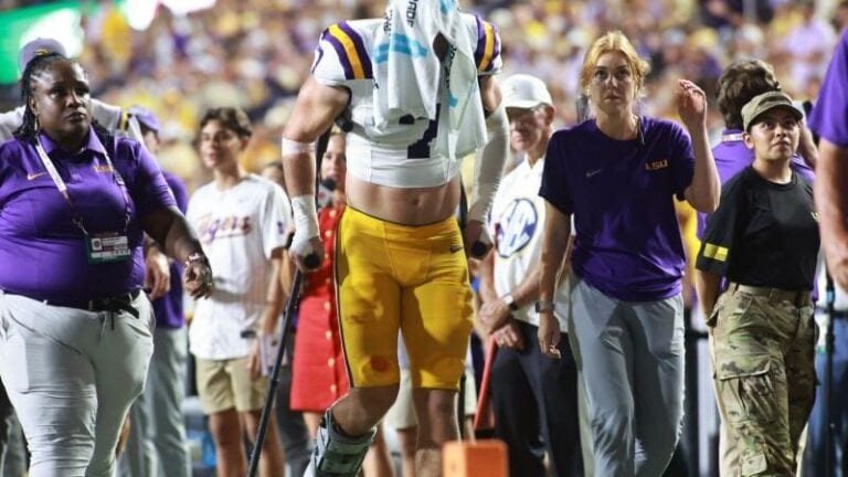 Whit Weeks, wearing yellow pants and a white towel on his head, walks on crutches after a leg injury, surrounded by staff and fans.