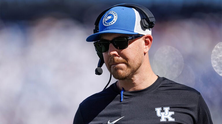 A coach wearing a Kentucky Wildcats cap, sunglasses, and a headset while providing sports analysis.