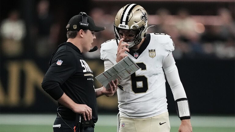 A New Orleans Saints coach talks to a quarterback holding a play sheet on the field.