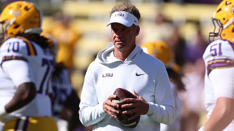 Lane Kiffin wears a white visor and hoodie while holding a football on the field between SEC players.