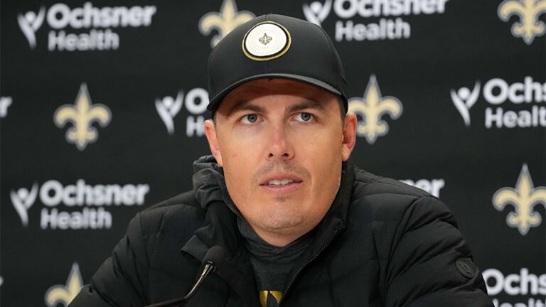 A man in a black Saints baseball cap with a gold fleur-de-lis logo speaks at a press conference.