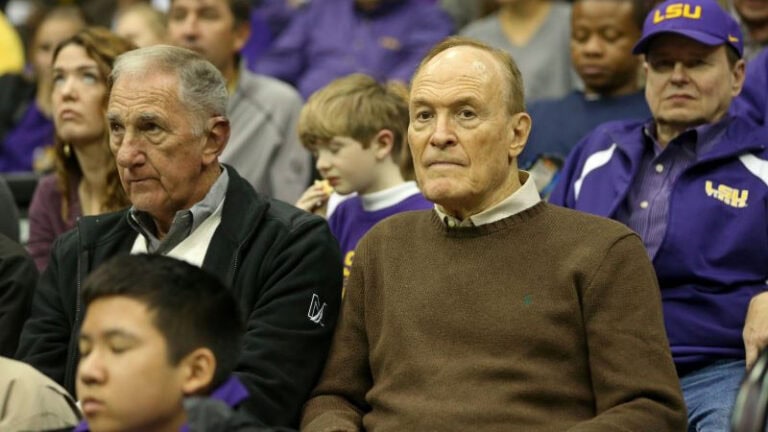 LSU coach Dale Brown sits with a man in a crowd at a sports event, one wearing a brown sweater and the other a black jacket.