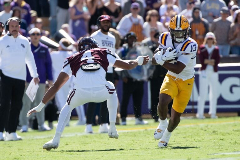 An LSU player in a white and gold uniform runs with the ball while a Mississippi State player attempts a tackle.