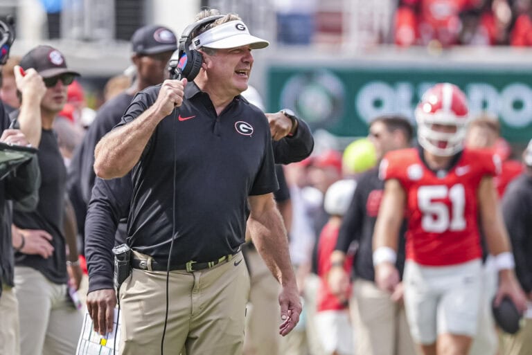 A college football coach in a visor and headset speaks into a microphone on the sidelines.