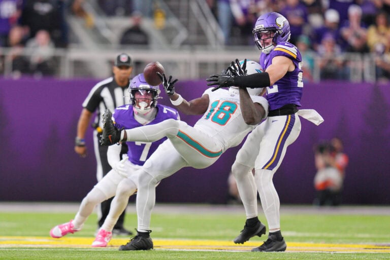 A Saints player in white reaches for a pass while being tackled by a Vikings defender in purple.