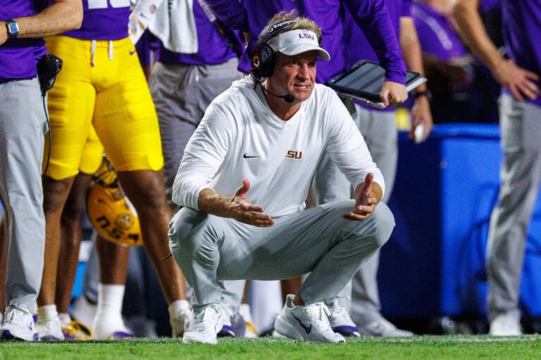 Lane Kiffin crouches on the field in a white hoodie and visor, signaling to his team.