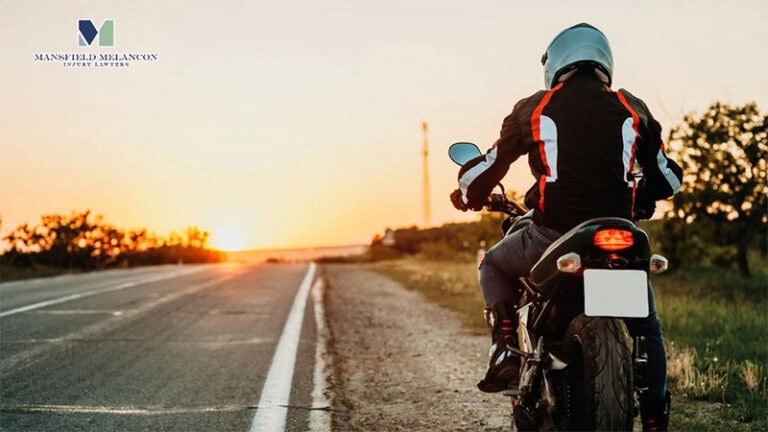 A motorcyclist in protective gear on a parked bike at sunset, featuring the Mansfield Melancon law firm logo.