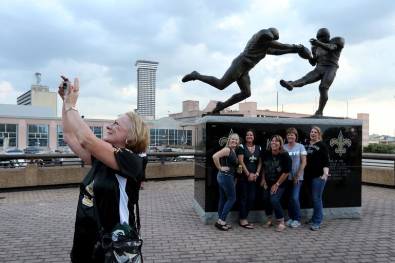 A woman takes a selfie with five friends in front of a bronze New Orleans Saints statue.