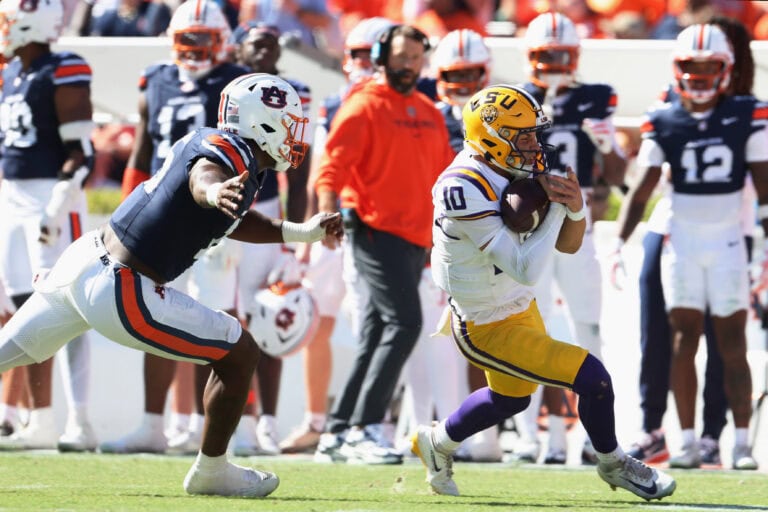 An LSU player in a white and gold uniform runs with the football while being chased by an opponent.